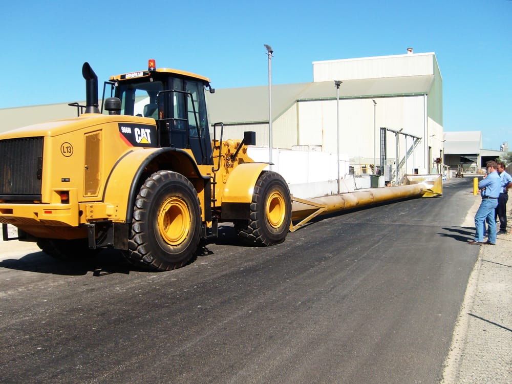 Cat 966 wheel loader carrying a long pipe on a sealed road beside a processing plant