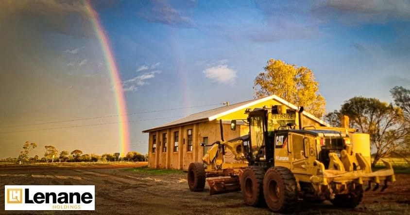 Cat motor grader parked beside a stone farm shed under a double rainbow at golden hour