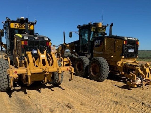Two Cat motor graders nose to nose on a gravel pad, one with an oversize sign