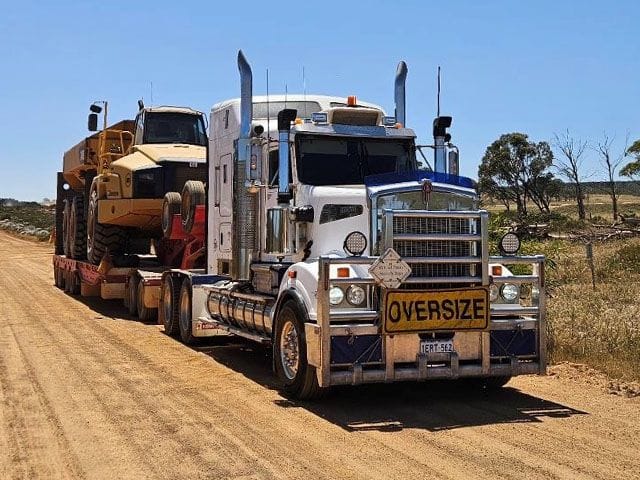 White Kenworth prime mover with an oversize sign hauling a Cat wheel loader on a float along a gravel road
