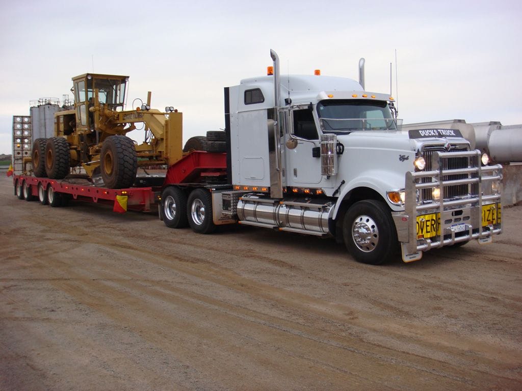 White Kenworth prime mover with an oversize sign carrying a Cat grader on a low loader along a dirt road