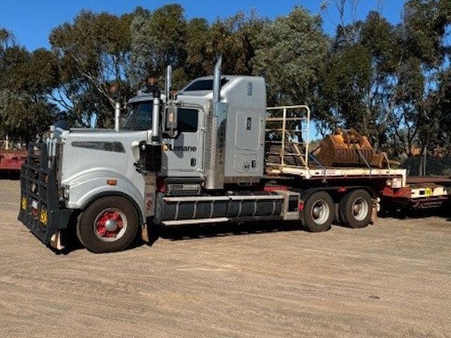 White Kenworth prime mover with a step deck trailer carrying plant, trees behind
