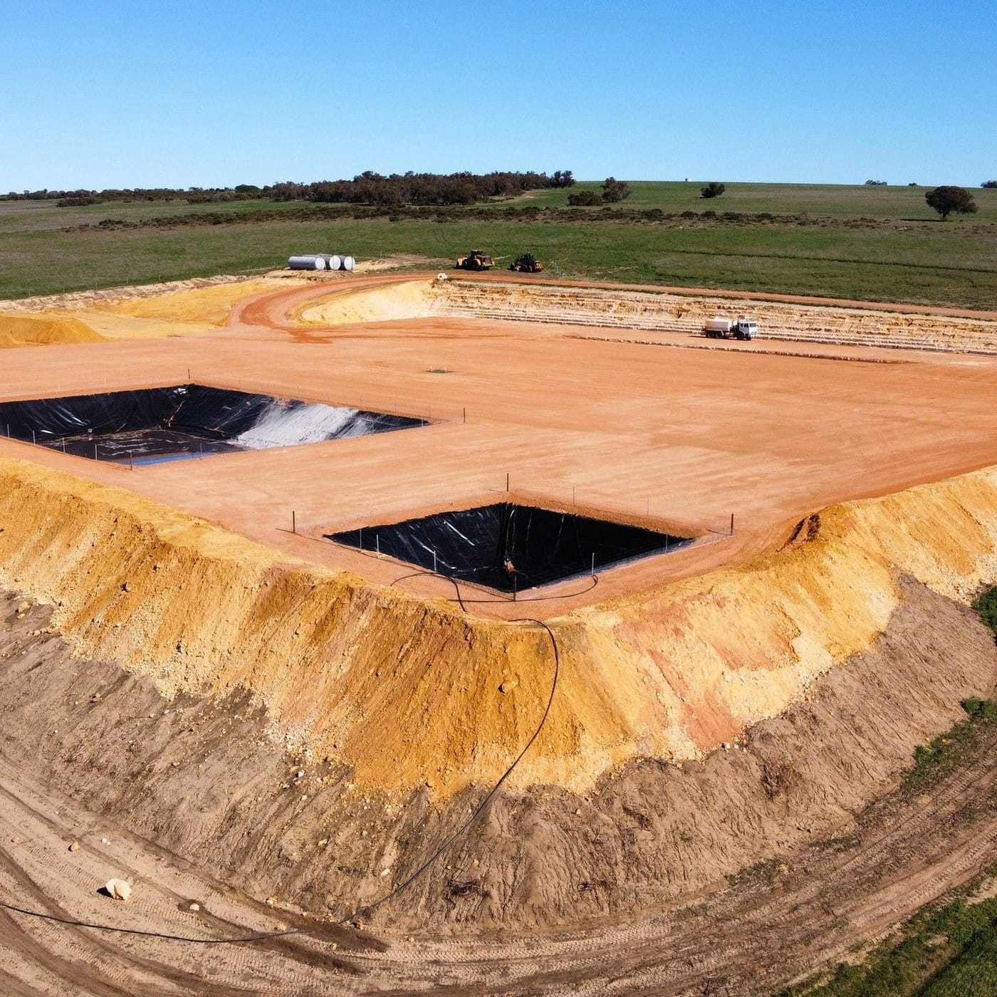 Drone view of lined sumps beside a red dirt pad, green paddocks around