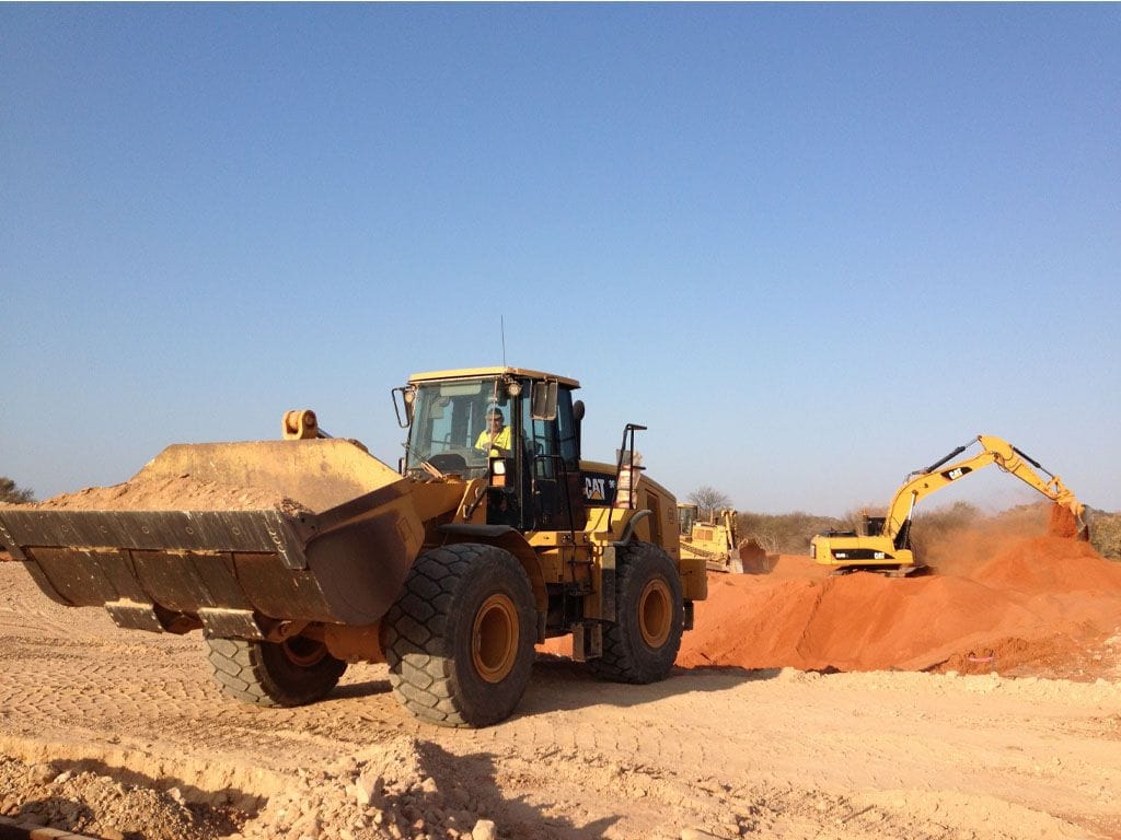 Cat wheel loader carrying a bucket of sand across red dirt, an excavator loading a tipper behind