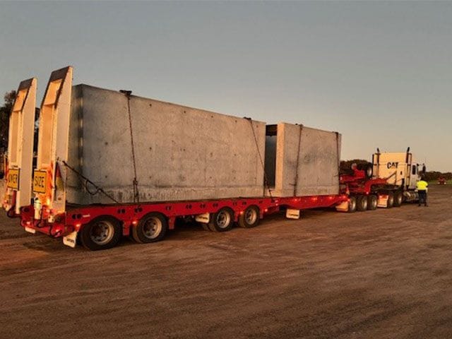 Red low loader trailer carrying large precast concrete units at dusk