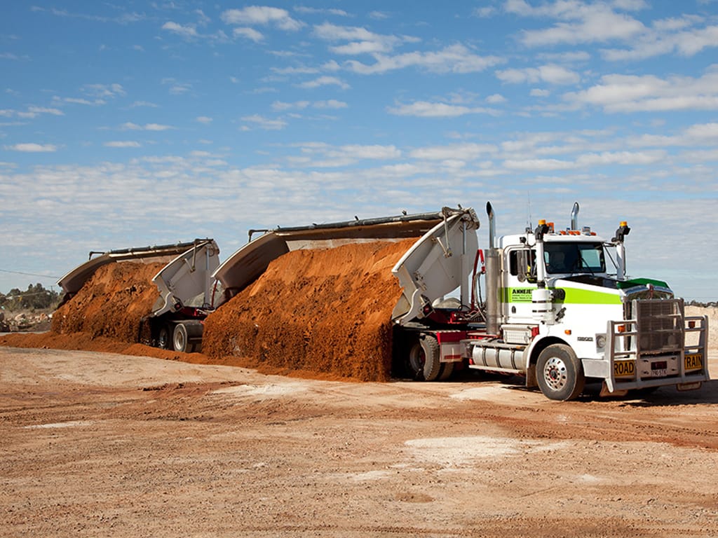 White Kenworth with side tippers tipping red gravel, bins raised under a blue sky