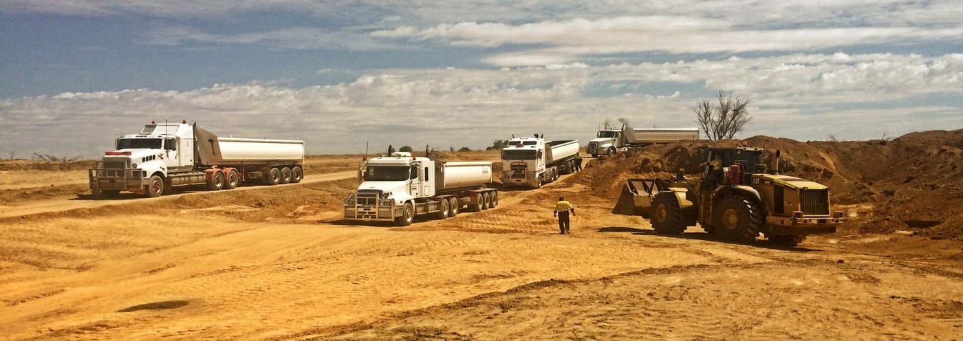 White tipper trucks and a loader on an open red dirt site under a blue sky