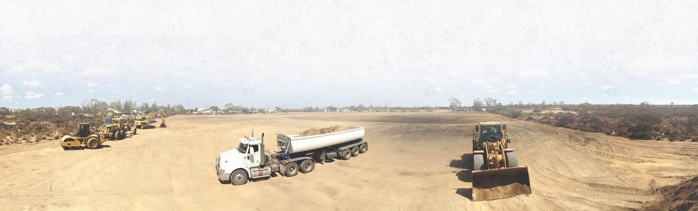 Wide panorama of a water tanker truck, a loader and tippers spread across a red dirt site